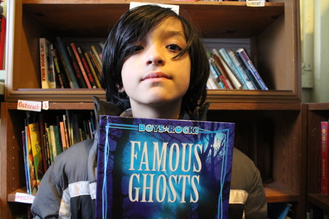 <p>Jos&eacute;, 10, holds up a book on ghosts. Behind him is a bookcase of books from The People&rsquo;s Library, which teacher Rachel Perveiler started on the back porch of her Little Village apartment.</p>
