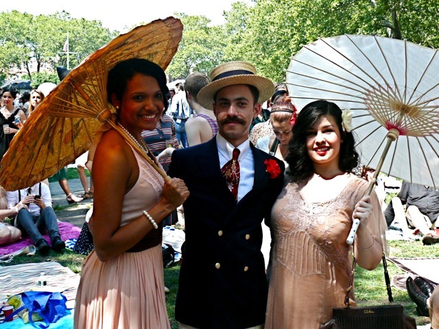 A gentleman in a double- breasted navy blue blazer and hard straw boater with two 30s-inspired ladies 