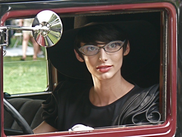 Posing in an antique car wearing a 30s style black ruffle shoulder dress a wide brimmed hat and Alain Mikili eyewear