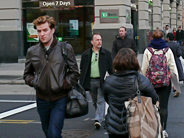 A chocolate brown bomber-influenced biker jacket with side-parted hair and a wind-blown roll, walking on Fifth Avenue. 