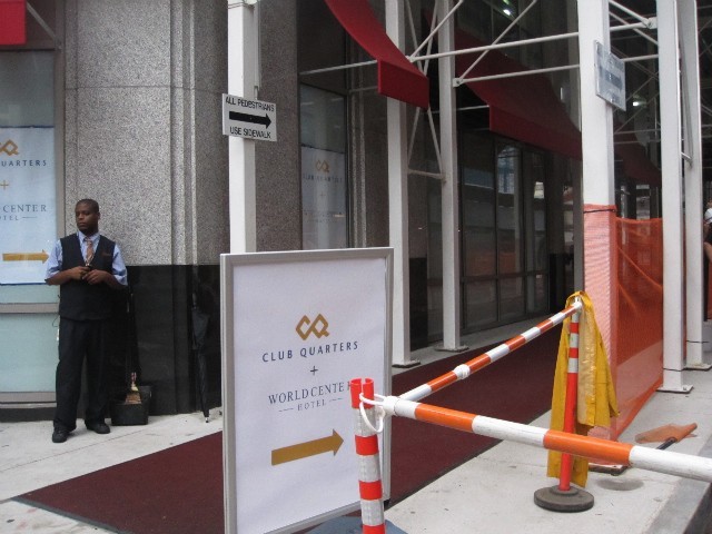 A bellhop waits to escort guests along a red carpet beneath a sidewalk shed to the hotel's entrance.